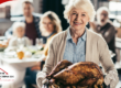 Older woman smiling while holding a large, golden roasted turkey in the kitchen, with a family gathered at the dining table in the background.