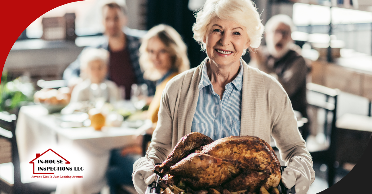 Older woman smiling while holding a large, golden roasted turkey in the kitchen, with a family gathered at the dining table in the background.